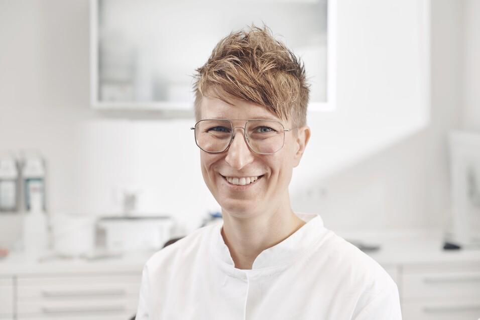 Portr&auml;t einer l&auml;chelnden Person mit Brille, die einen wei&szlig;en Kittel in einem modernen, hellen Raum tr&auml;gt.