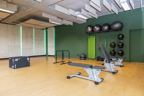 Fitnessraum mit Hanteln, Trainingsbank, Klimmzugstangen und einer schwarz-grauen Plyobox auf einem Holzfußboden.