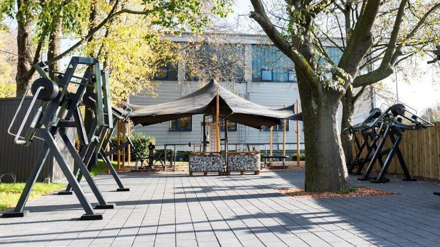 Outdoor-Fitnessbereich mit schwarzen Trainingsgeräten, umgeben von Bäumen und einem Gebäude im Hintergrund.