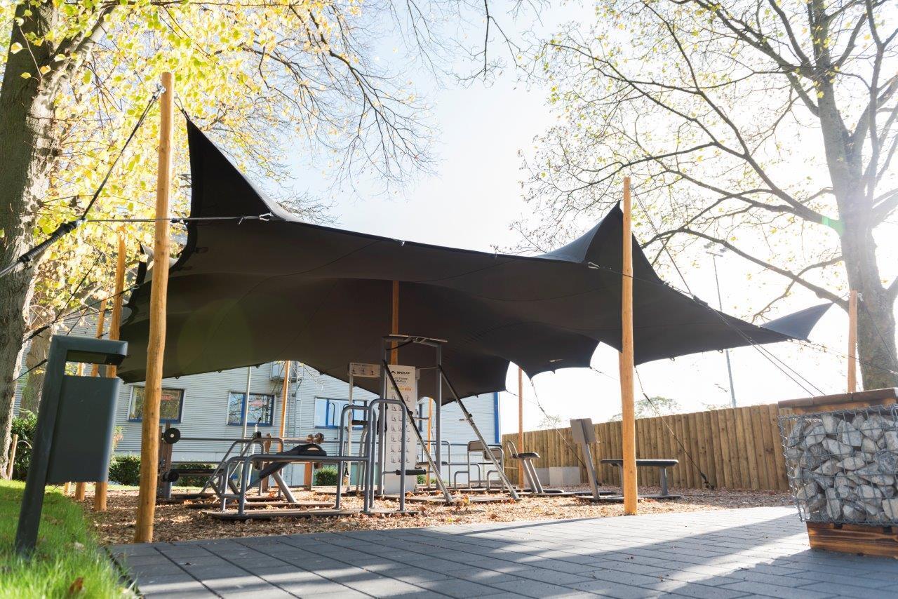 Schattenspendendes Sonnensegel &uuml;ber einem Spielplatz mit mehreren Ger&auml;ten in einem begr&uuml;nten Au&szlig;enbereich.