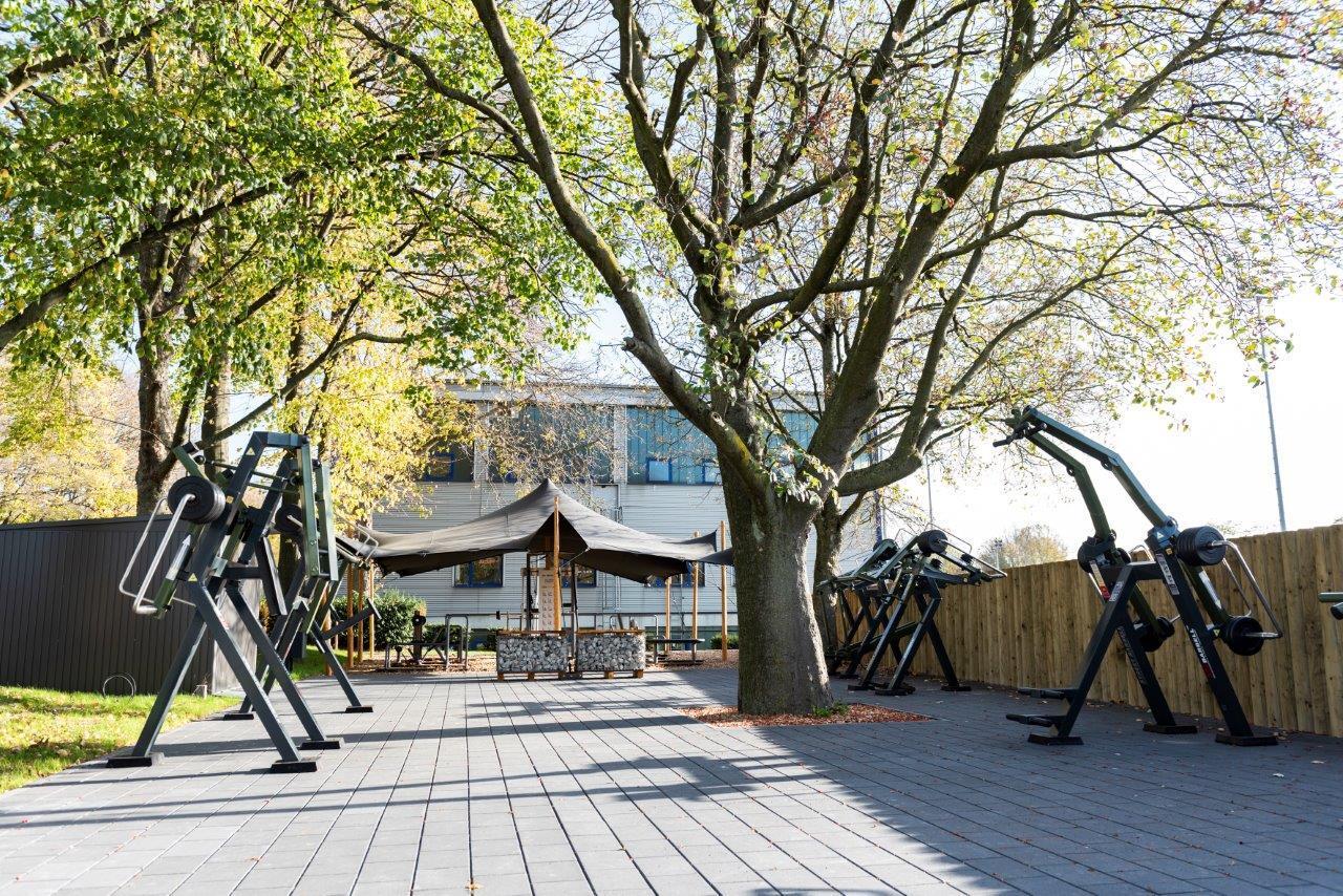 Outdoor-Fitnessbereich mit schwarzen Trainingsger&auml;ten, umgeben von B&auml;umen und einem Geb&auml;ude im Hintergrund.