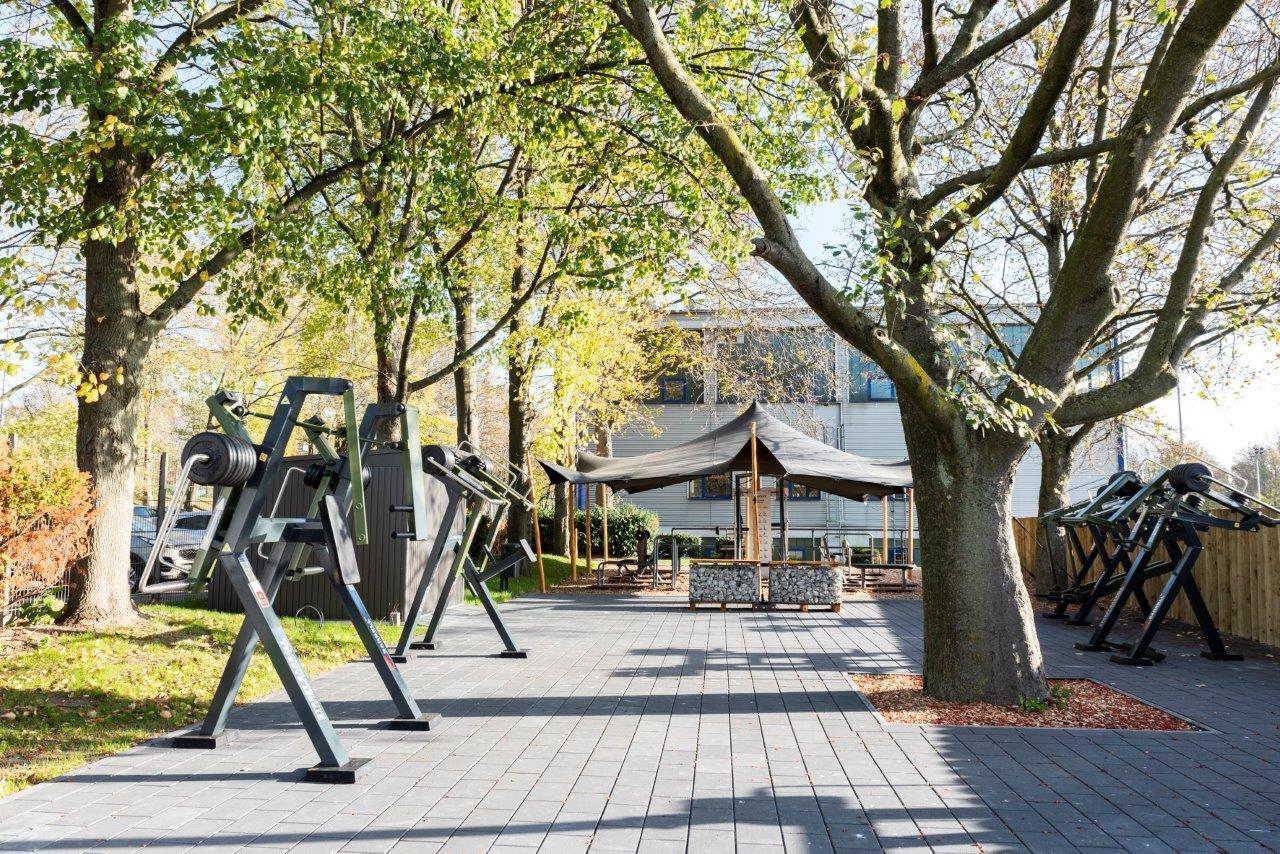 Freiluftfitnessbereich mit Trainingsger&auml;ten, umgeben von B&auml;umen und einer Sitzgelegenheit unter einem Pavillon.