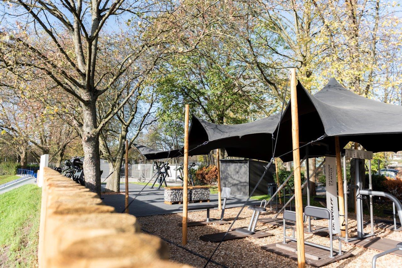 Outdoor-Fitnessbereich mit schwarzen Sonnenschutzmarkisen und Trainingsger&auml;ten, umgeben von B&auml;umen und einem Zaun.