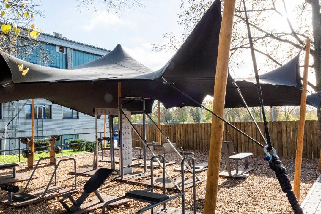 Freiluft-Fitnessbereich mit schwarzen Sonnensegeln und verschiedenen Trainingsger&auml;ten auf einem Holzschnitzelboden.
