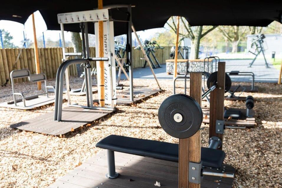 Outdoor-Fitnessbereich mit verschiedenen Geräten und Holzplattformen, teilweise beschattet durch ein Zeltdach.