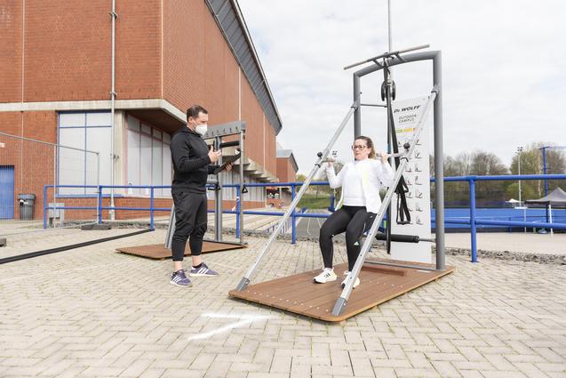 Frau trainiert an einem multifunktionalen Fitnessger&auml;t im Freien, w&auml;hrend ein Trainer sie anleitet.