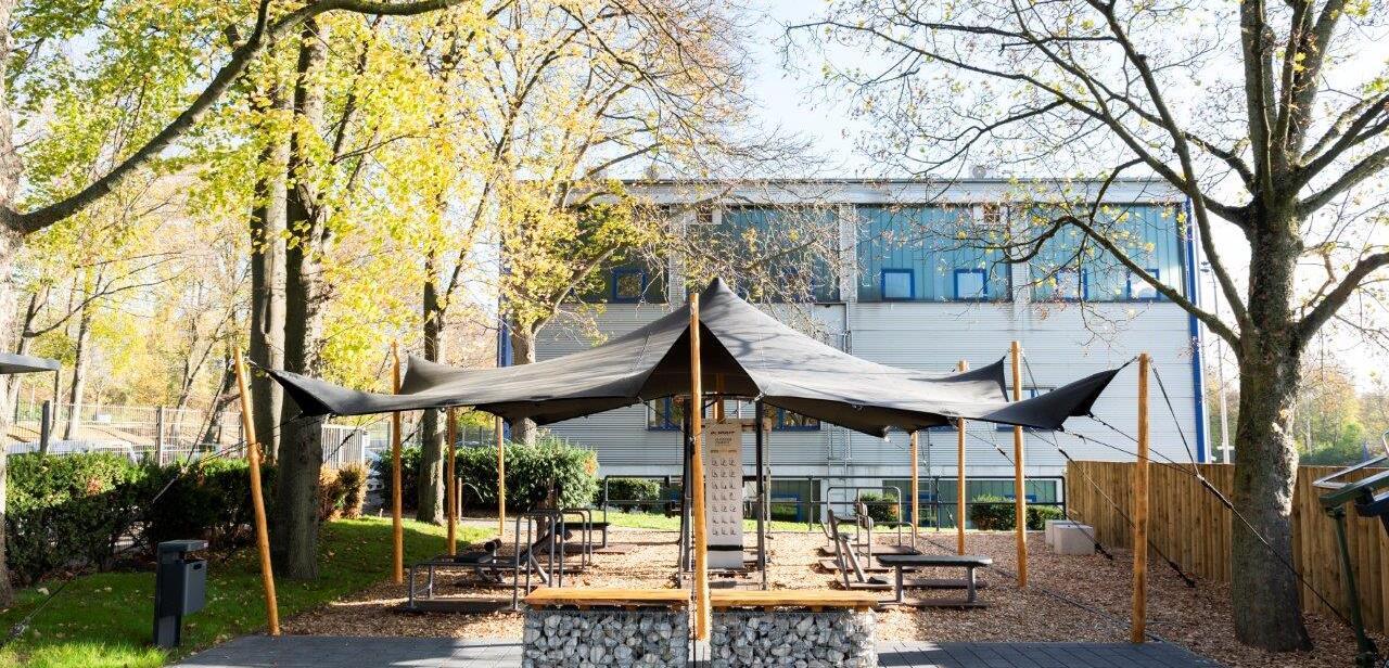 Überdachte Sitzarea mit Holzbänken und Tisch, umgeben von Bäumen und einem modernen Gebäude im Hintergrund.
