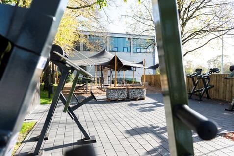 Blick auf einen Outdoor-Fitnessbereich mit Ger&auml;ten und einem Pavillon im Hintergrund, umgeben von B&auml;umen.
