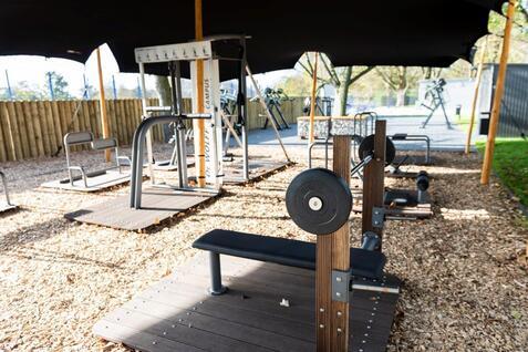 Outdoor-Fitnessbereich mit verschiedenen Ger&auml;ten und Holzplattformen, teilweise beschattet durch ein Zeltdach.