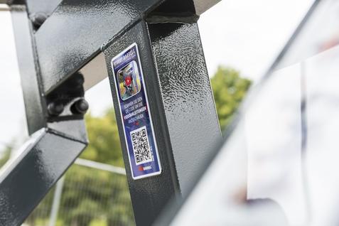 Nahaufnahme eines QR-Codes und einer Informationskarte an einem grauen Metallger&uuml;st. Hintergrund mit verschwommenen B&auml;umen.