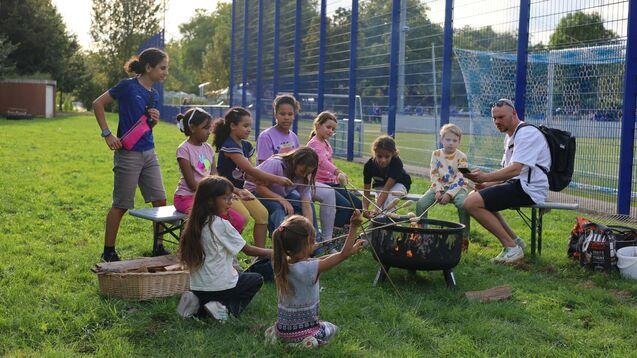 Gruppe von Kindern und zwei Erwachsenen am Lagerfeuer, w&auml;hrend sie Stockbrot auf einer Wiese zubereiten.