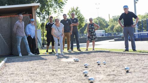 Mehrere Personen stehen und sitzen an einem Boule-Feld im Freien. Im Vordergrund wirft eine ältere Frau konzentriert eine Boule-Kugel, während die anderen gespannt zusehen. Die Szene spielt sich bei sonnigem Wetter auf einer Sportanlage ab.