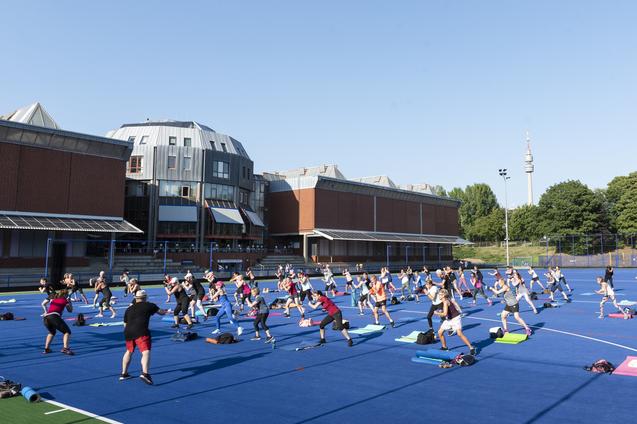 Gruppe von Menschen bei einem Outdoor-Workout auf einer blauen Sportfl&auml;che vor einem modernen Geb&auml;ude.