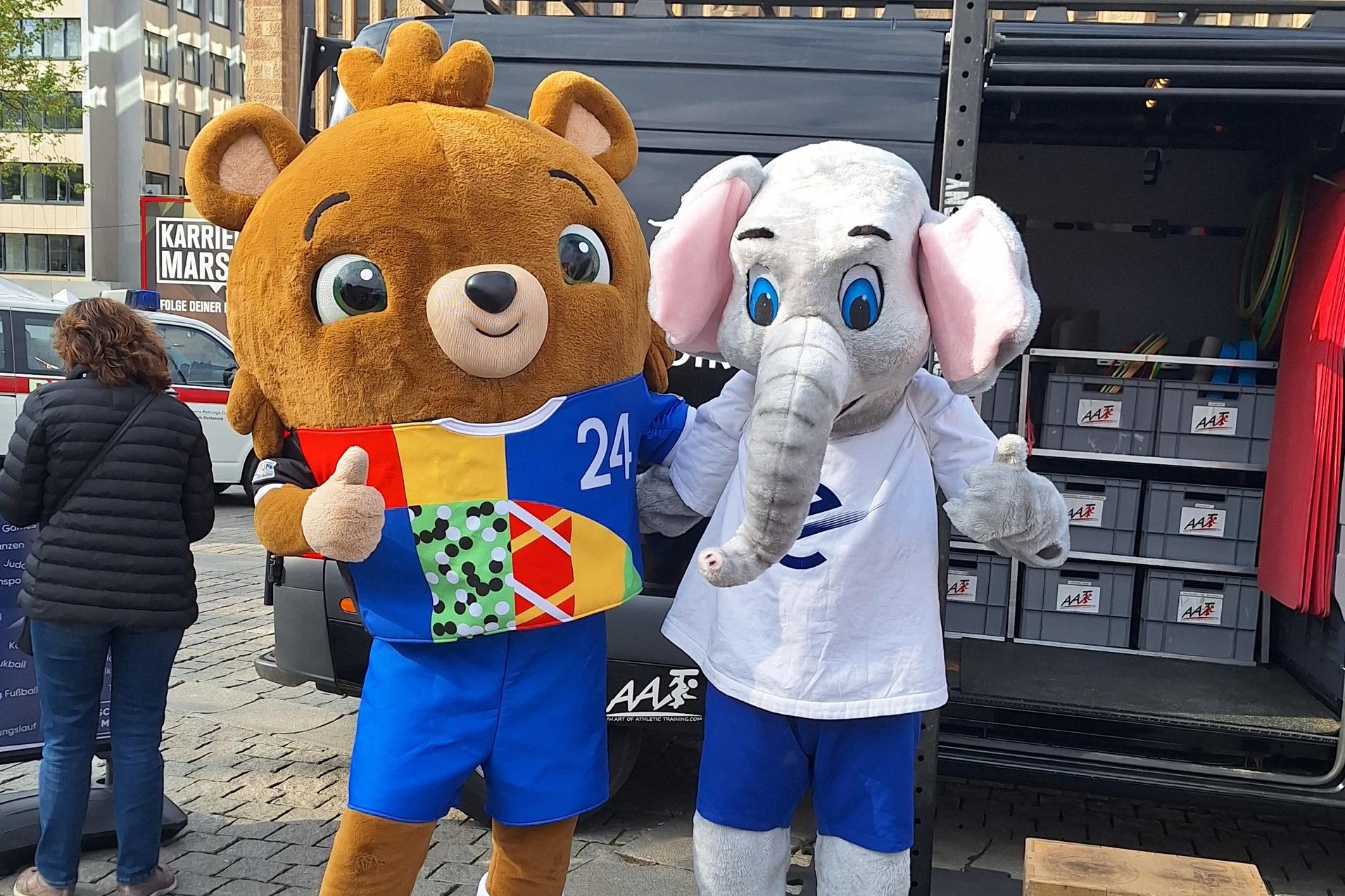 Zwei Maskottchen, ein B&auml;r mit buntem Shirt und ein Elefant, stehen vor einem Lieferwagen auf einem Platz.