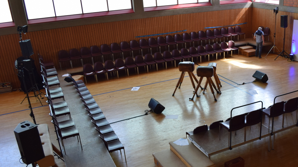 Blick auf eine leere Halle mit Stuhlreihen, einer B&uuml;hne und Lautsprechern f&uuml;r eine bevorstehende Veranstaltung.