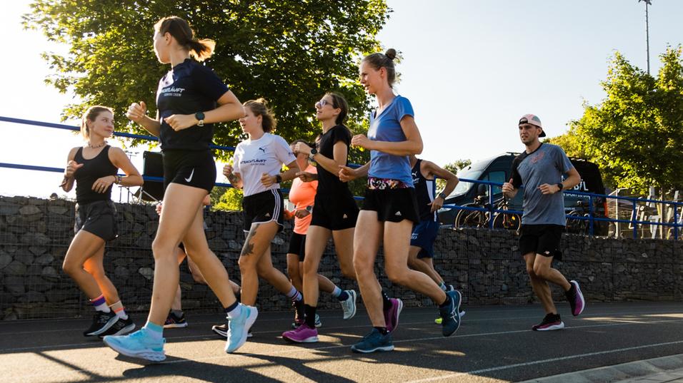 Gruppe von acht Personen joggt auf einem Gehweg, umgeben von Bäumen und Fahrrädern im Hintergrund.