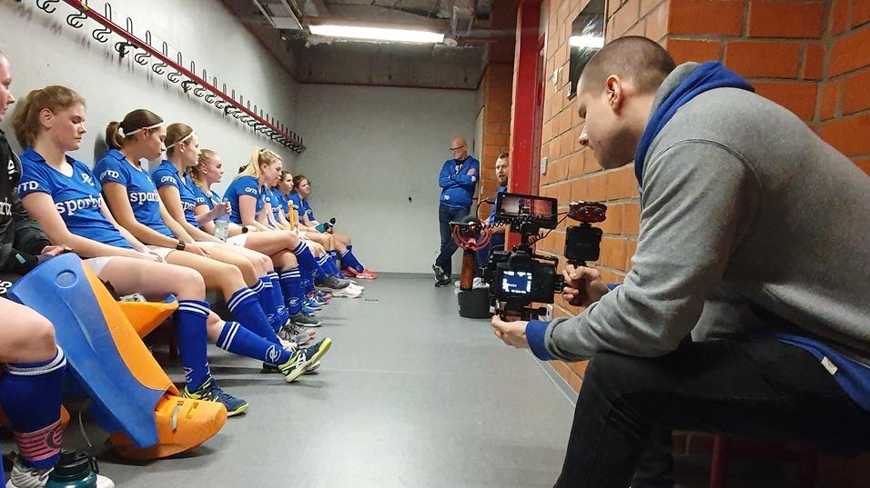 Frauenfußballmannschaft in blauen Trikots sitzt in der Umkleidekabine, während ein Filmer sie dokumentiert.