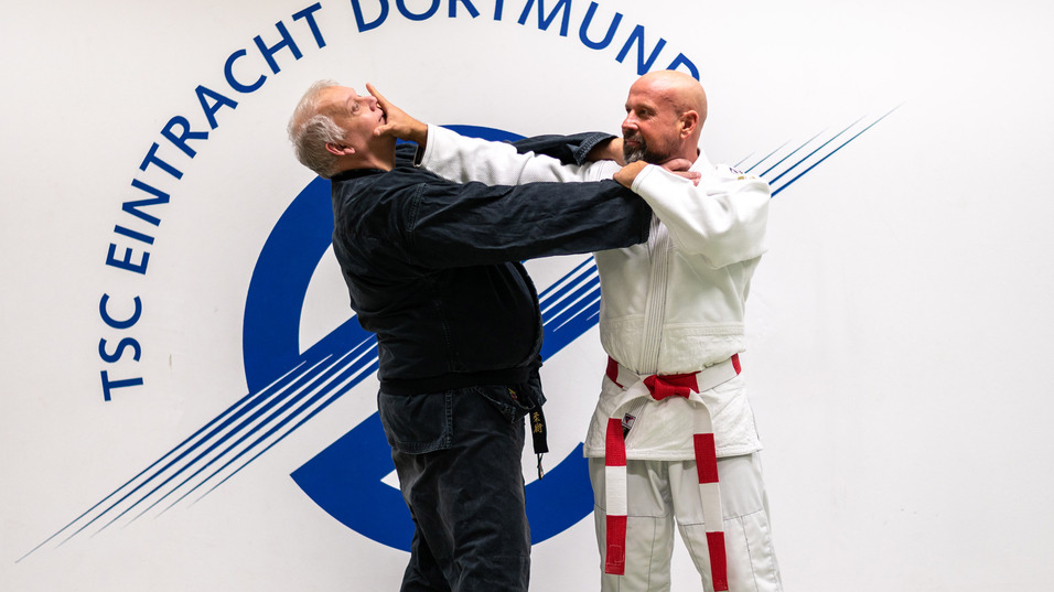 Zwei Judoka in Aktion, einer mit schwarzem, der andere mit wei&szlig;em Gi, vor einem Wandbanner des TSC Eintracht Dortmund.