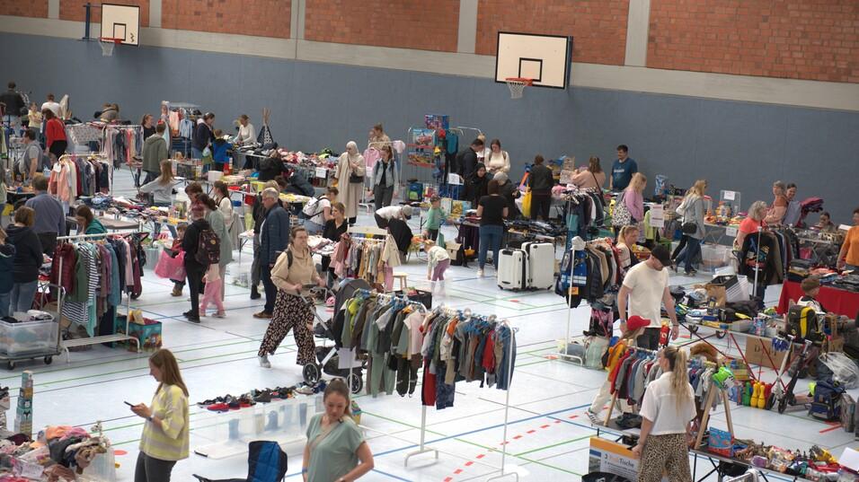 Markt mit zahlreichen St&auml;nden und Menschen, die Kleidung und Haushaltswaren in einer Sporthalle betrachten und kaufen.