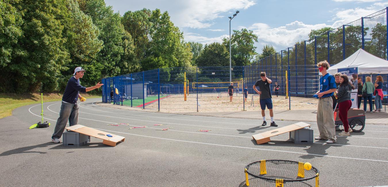 Zwei Gruppen spielen Spiele auf einer sportlichen Anlage mit Sandplatz und öffentlichen Einrichtungen im Hintergrund.