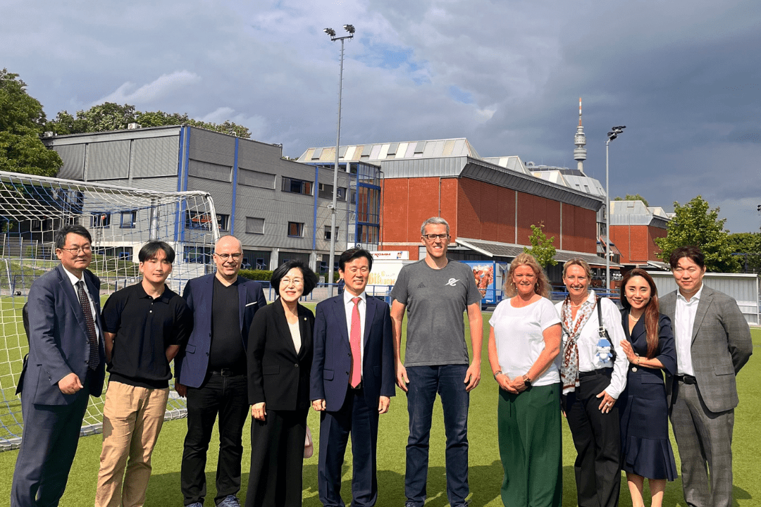 Gruppe von zehn Personen auf einem Sportplatz, hinter ihnen moderne Gebäude und ein Fußballtor, bei bewölktem Himmel.