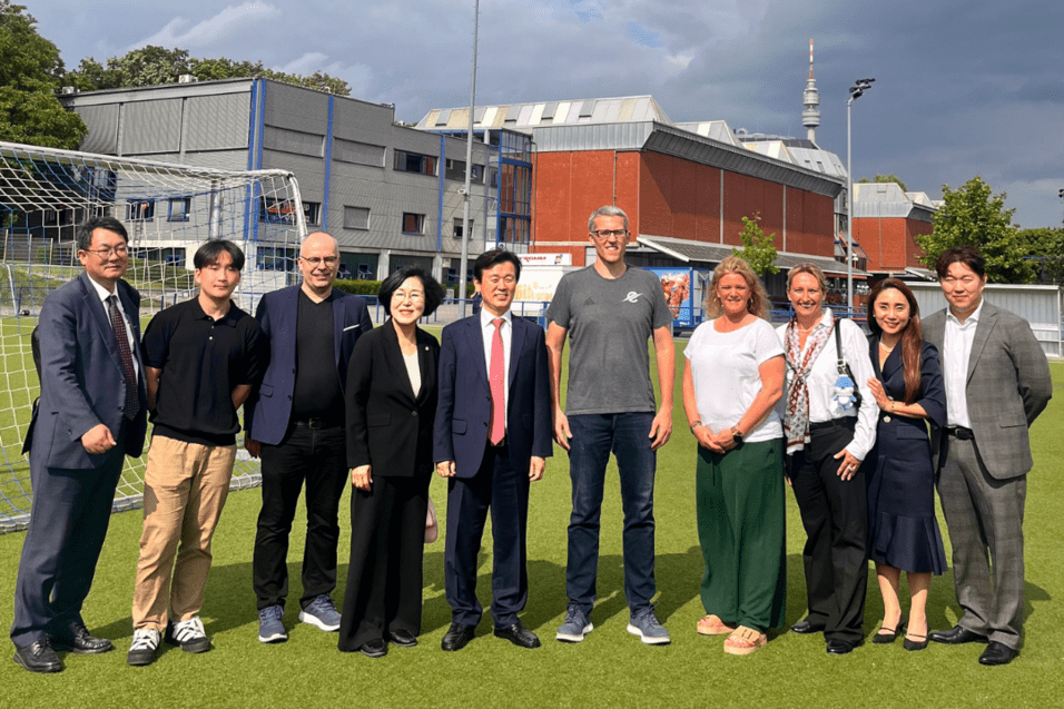 Gruppe von zehn Personen auf einem Sportplatz, hinter ihnen moderne Geb&auml;ude und ein Fu&szlig;balltor, bei bew&ouml;lktem Himmel.