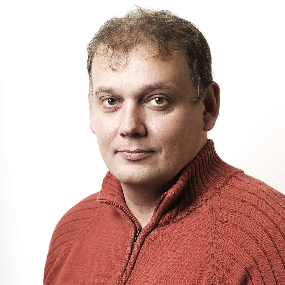 Porträt eines Mannes mit kurzen Haaren, der ein rotes, hochgeschlossenes Sweatshirt trägt, vor einem weißen Hintergrund.