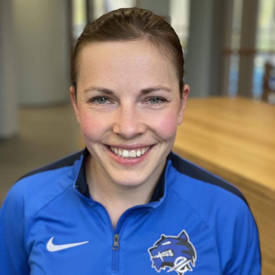 L&auml;chelnde Frau mit kurzen Haaren tr&auml;gt einen blauen Sportpullover mit Logo. Helle Innenr&auml;ume im Hintergrund.