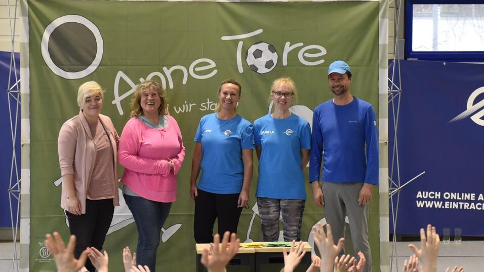 Gruppenszene mit fünf Personen vor einer grünen Wand, während Kinder im Vordergrund applaudieren und die Hände heben.