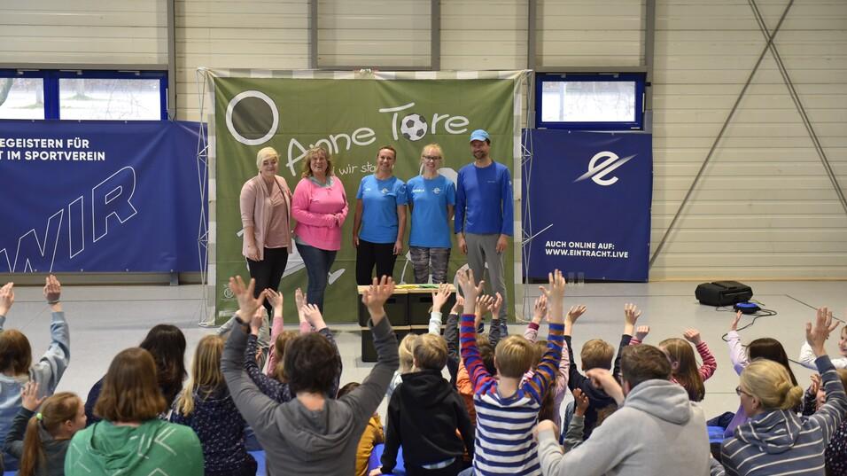 Gruppenszene mit fünf Personen vor einer grünen Wand, während Kinder im Vordergrund applaudieren und die Hände heben.