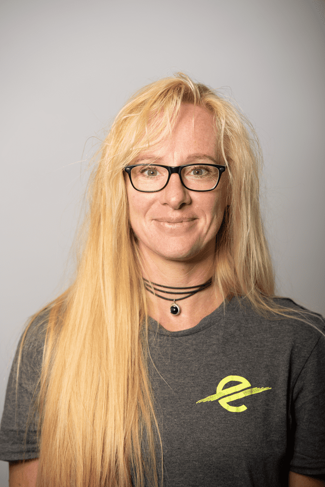 Porträt einer lächelnden Frau mit langen blonden Haaren, Brille und grauem T-Shirt mit gelbem Logo. Hintergrund grau.