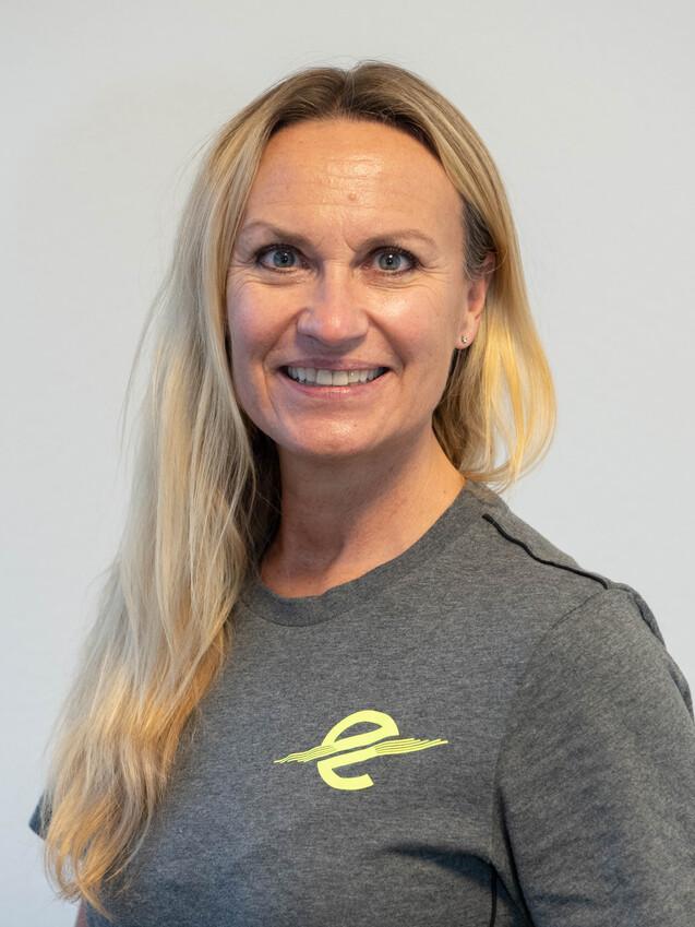 Porträt einer lächelnden Frau mit langen blonden Haaren, die ein graues T-Shirt mit gelbem Logo trägt.