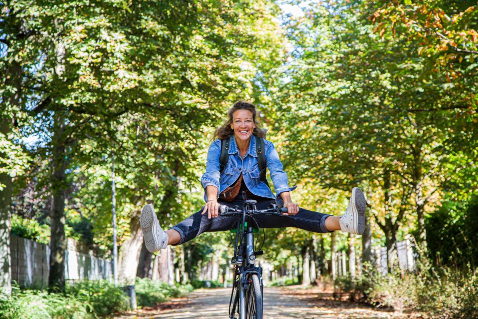 Junge Frau fährt auf einem Fahrrad, mit gespreizten Beinen, lächelnd in einer Allee mit herbstlichen Bäumen.