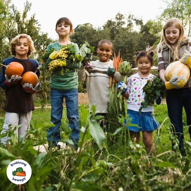 Fünf Kinder stehen im Garten und halten verschiedene Gemüse und Früchte, umgeben von Pflanzen und Grün.