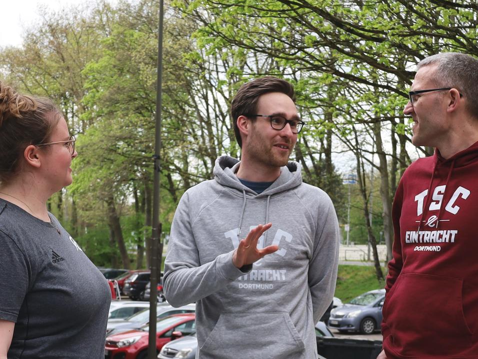 Drei Personen unterhalten sich im Freien, umgeben von Bäumen und parkenden Autos.