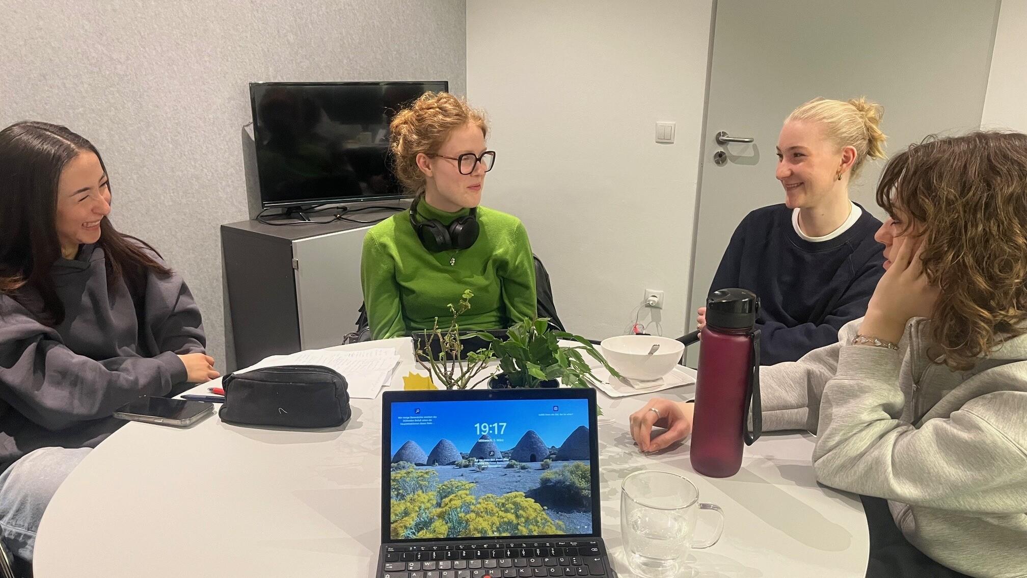 Vier Frauen sitzen an einem runden Tisch, w&auml;hrend ein Laptop und Pflanzen im Vordergrund sichtbar sind.