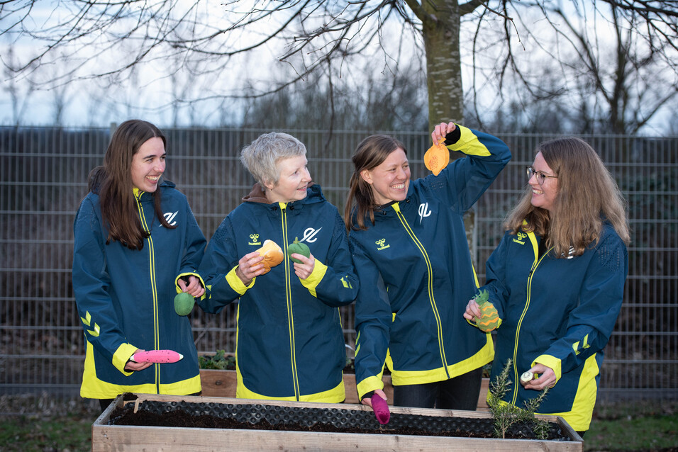Vier Frauen in blauen Jacken stehen um ein Hochbeet und halten buntes Gem&uuml;se in den H&auml;nden.