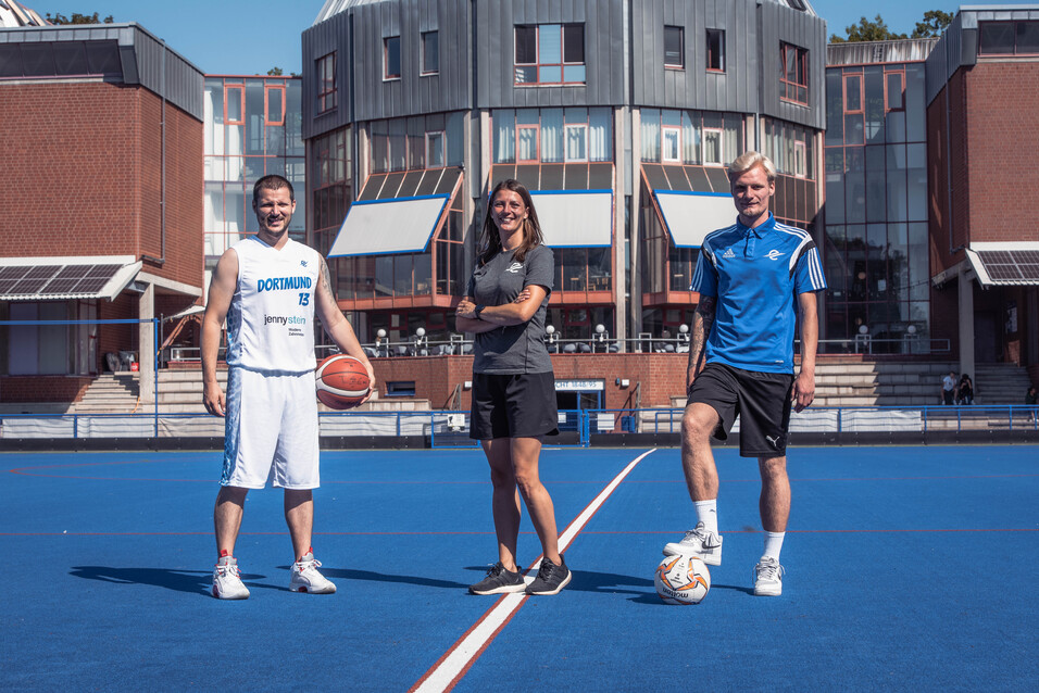 Drei Sportler stehen auf einem blauen Sportplatz: ein Basketballspieler mit Ball, eine Trainerin und ein Fu&szlig;ballspieler.