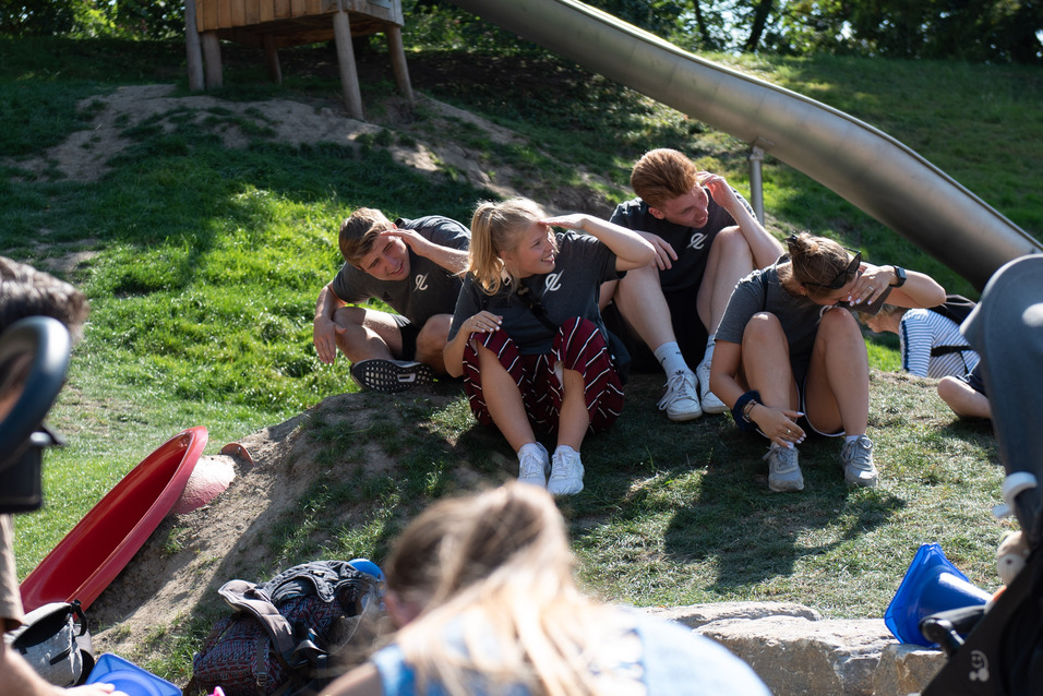 Gruppe von Kindern sitzt am Hang neben einer Rutsche, w&auml;hrend Erwachsene mit Kinderwagen im Vordergrund besch&auml;ftigt sind.