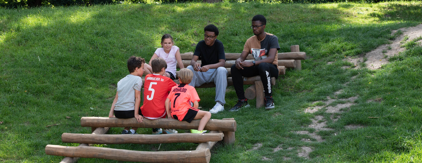 Sechs Kinder sitzen auf Holzstufen auf einem gr&uuml;nen H&uuml;gel und unterhalten sich im Freien.