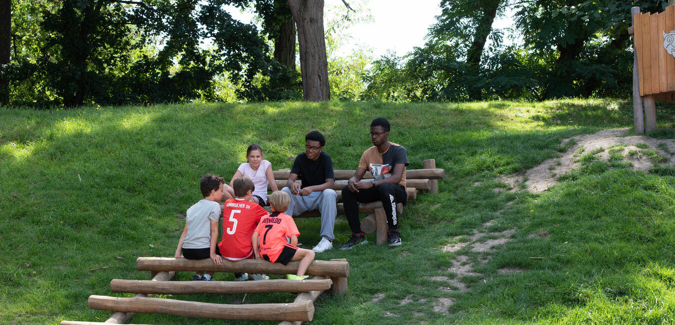 Sechs Kinder sitzen auf Holzstufen auf einem gr&uuml;nen H&uuml;gel und unterhalten sich im Freien.