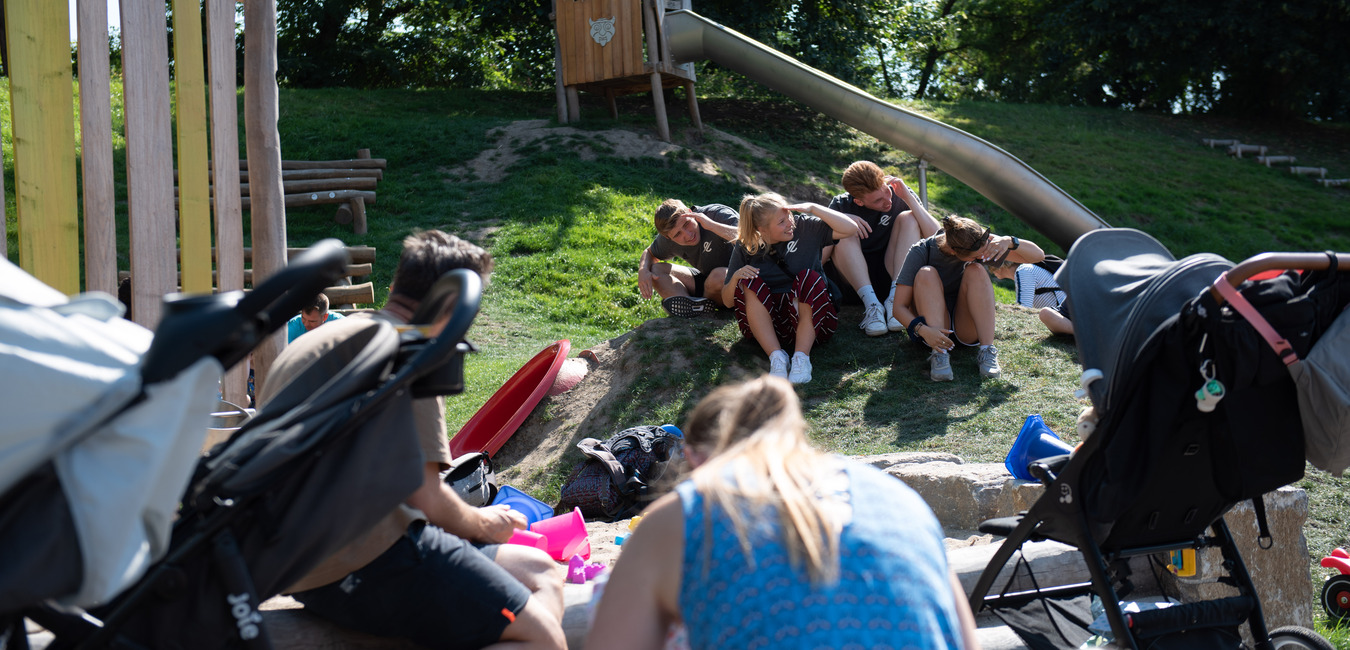 Gruppe von Kindern sitzt am Hang neben einer Rutsche, w&auml;hrend Erwachsene mit Kinderwagen im Vordergrund besch&auml;ftigt sind.