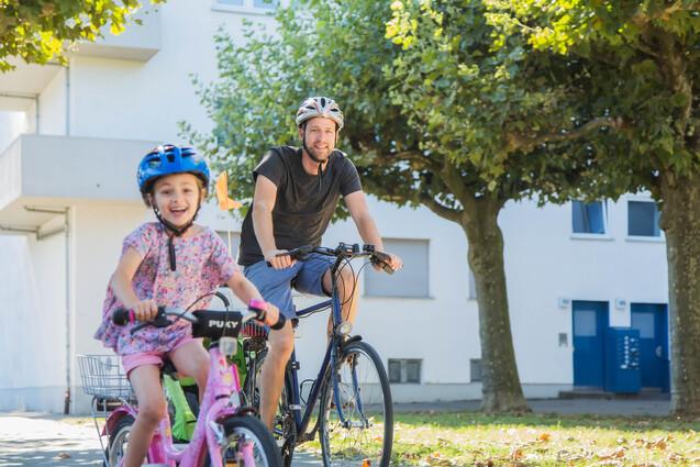 Vater und Tochter fahren gemeinsam mit Fahrrädern auf einem sonnigen Weg, umgeben von Bäumen und Wohngebäuden.
