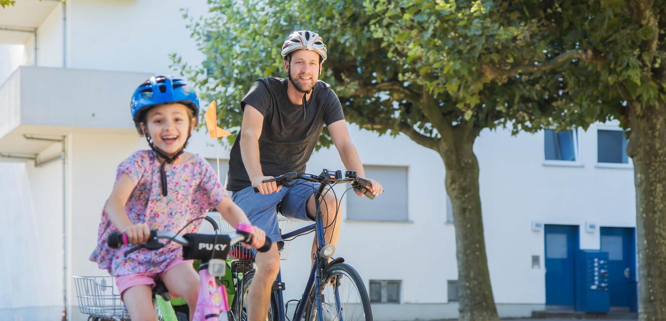 Vater und Tochter fahren gemeinsam mit Fahrrädern auf einem sonnigen Weg, umgeben von Bäumen und Wohngebäuden.