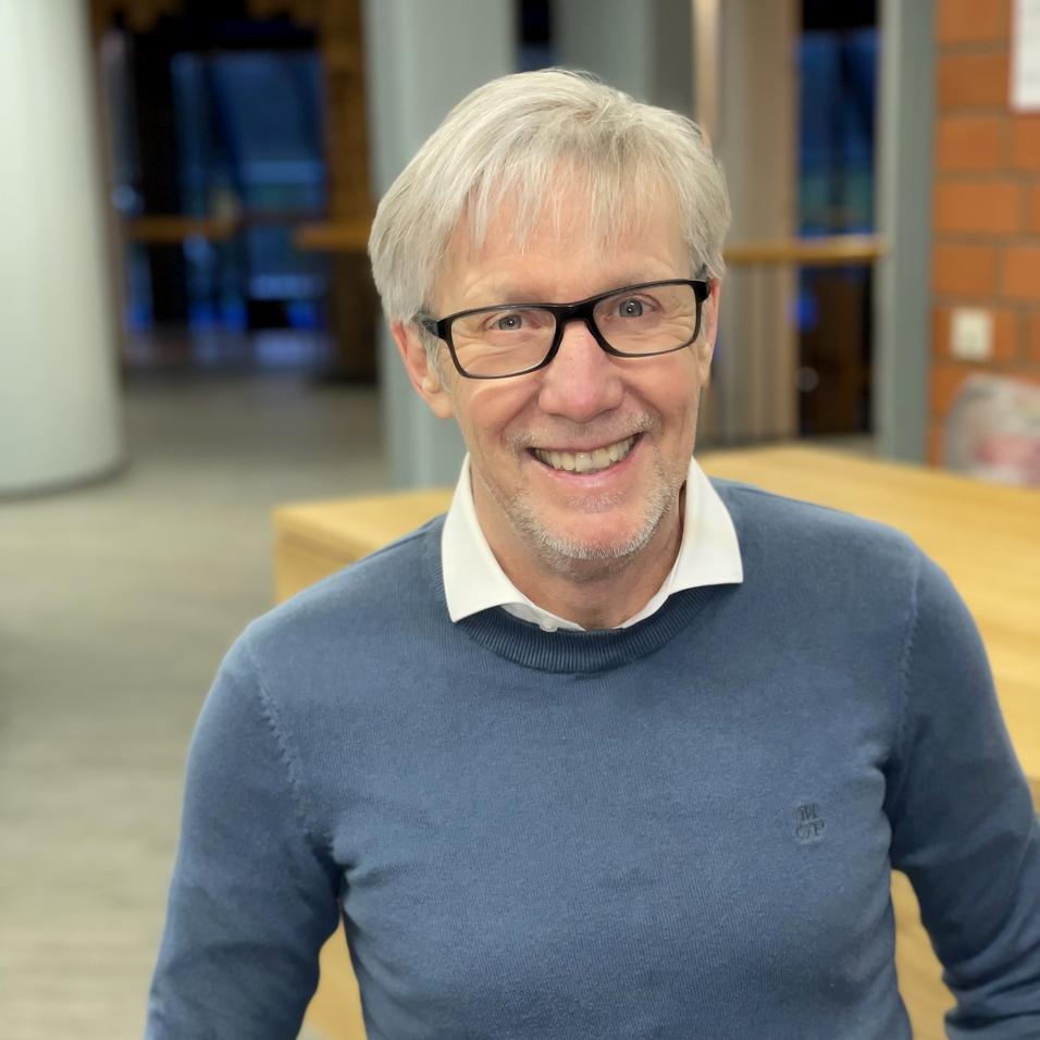 L&auml;chelnder Mann mit Brille und grauen Haaren sitzt in einem modernen Raum, tr&auml;gt einen blauen Pullover &uuml;ber einem wei&szlig;en Hemd.