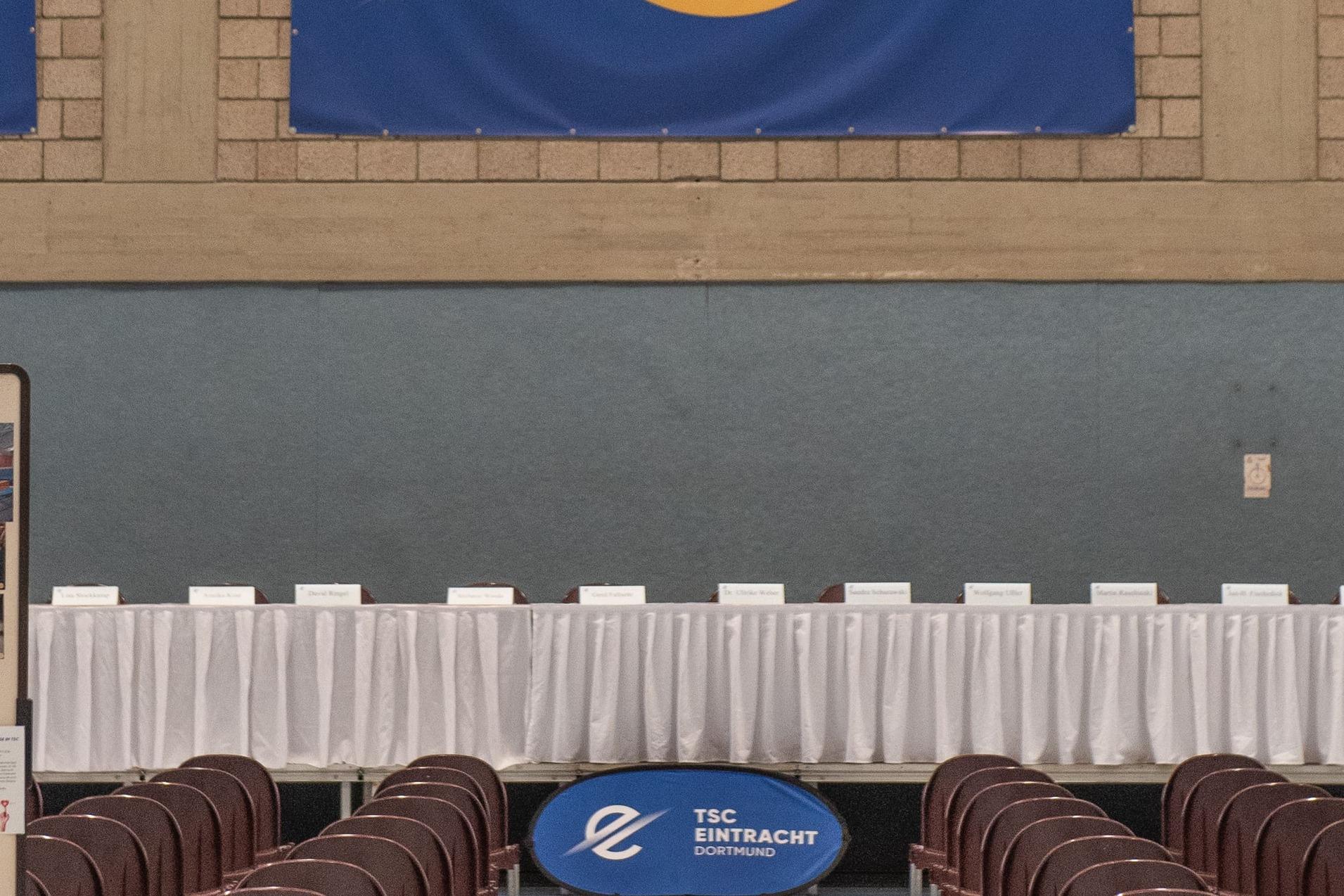 Innenansicht einer Sporthalle mit blauen Banner, Stuhlreihen und einem gedeckten Tisch am Ende.