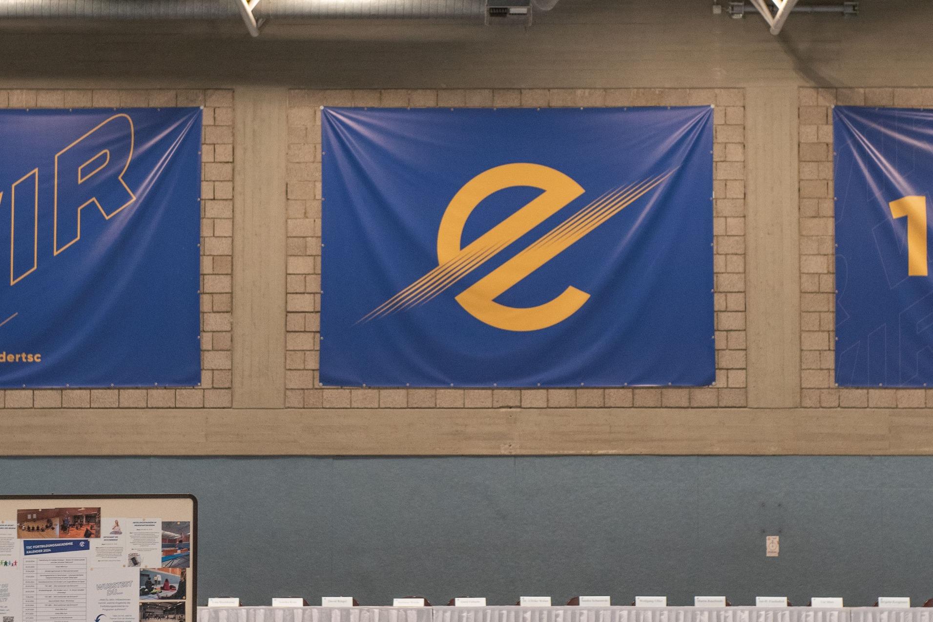 Innenansicht einer Sporthalle mit blauen Banner, Stuhlreihen und einem gedeckten Tisch am Ende.
