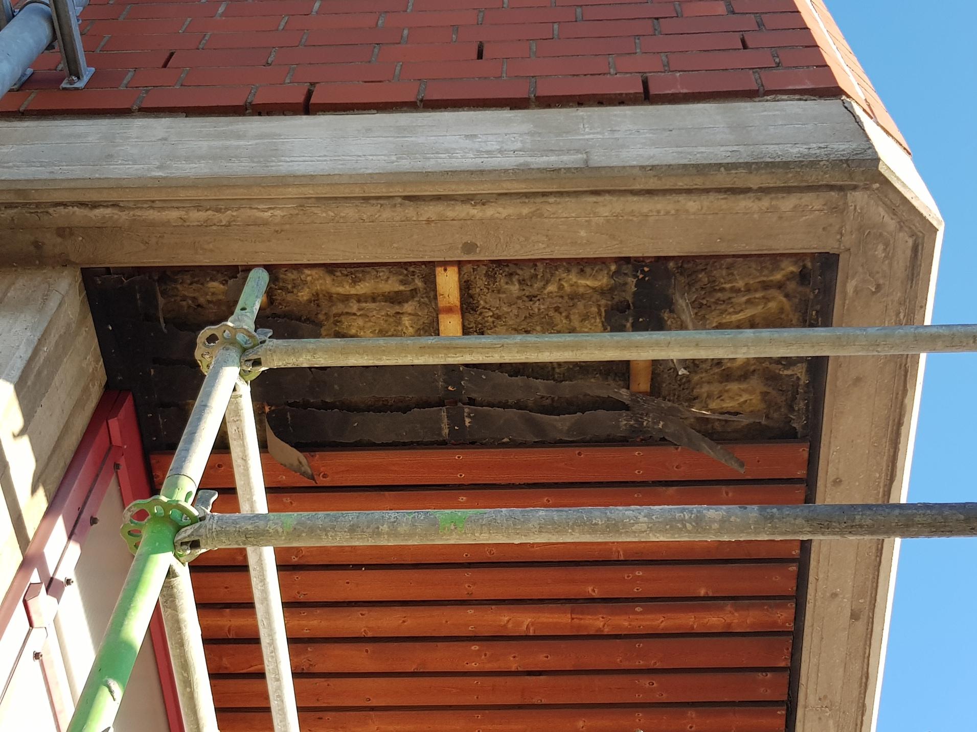 Blick auf eine unvollendete Geb&auml;udeverkleidung mit Holzrahmen und sichtbarem D&auml;mmmaterial unter einem Ger&uuml;st.