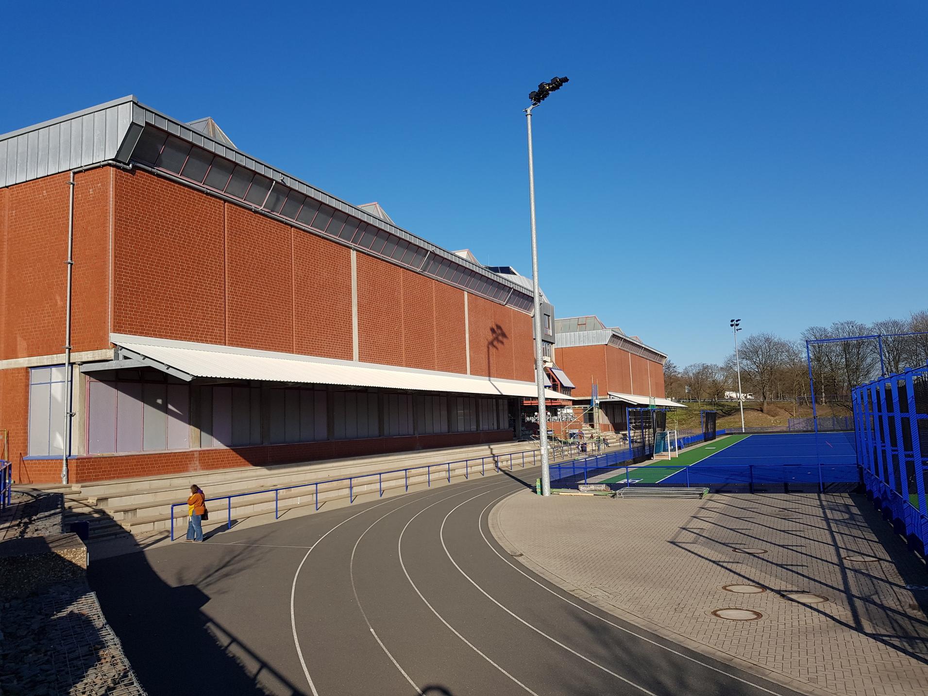Blick auf ein Sportgeb&auml;ude mit Laufbahn und Hockeyfeld, blauer Himmel im Hintergrund.