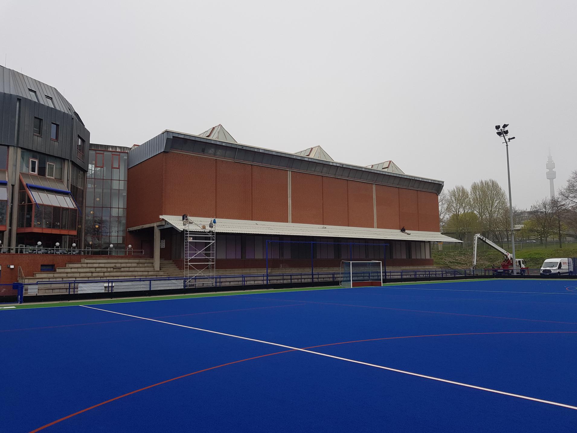 Modernes Sportgeb&auml;ude entlang eines blauen Hockeyfeldes, umgeben von Nebel und Vegetation. Im Hintergrund ein Fernsehturm.